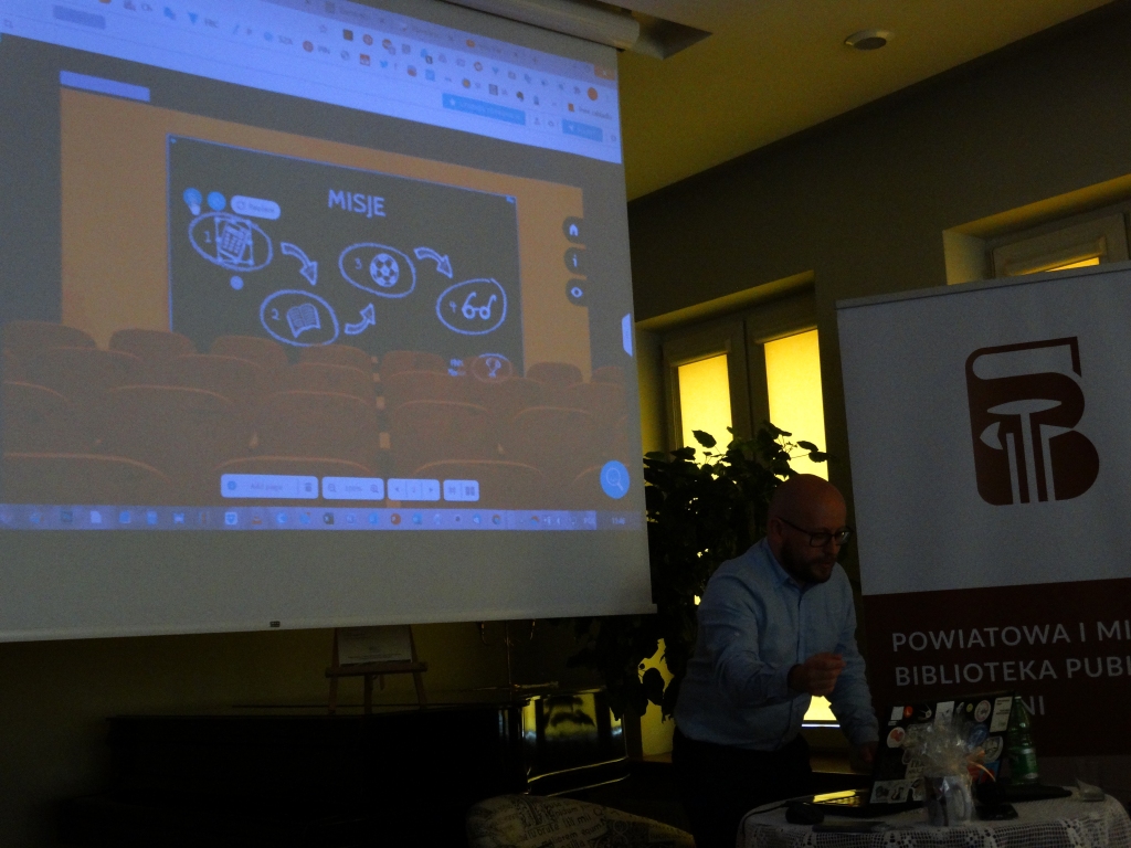 Siedzacy przy stoliku Karol Baranowski na tle ekranu z prezentacją multimedialną