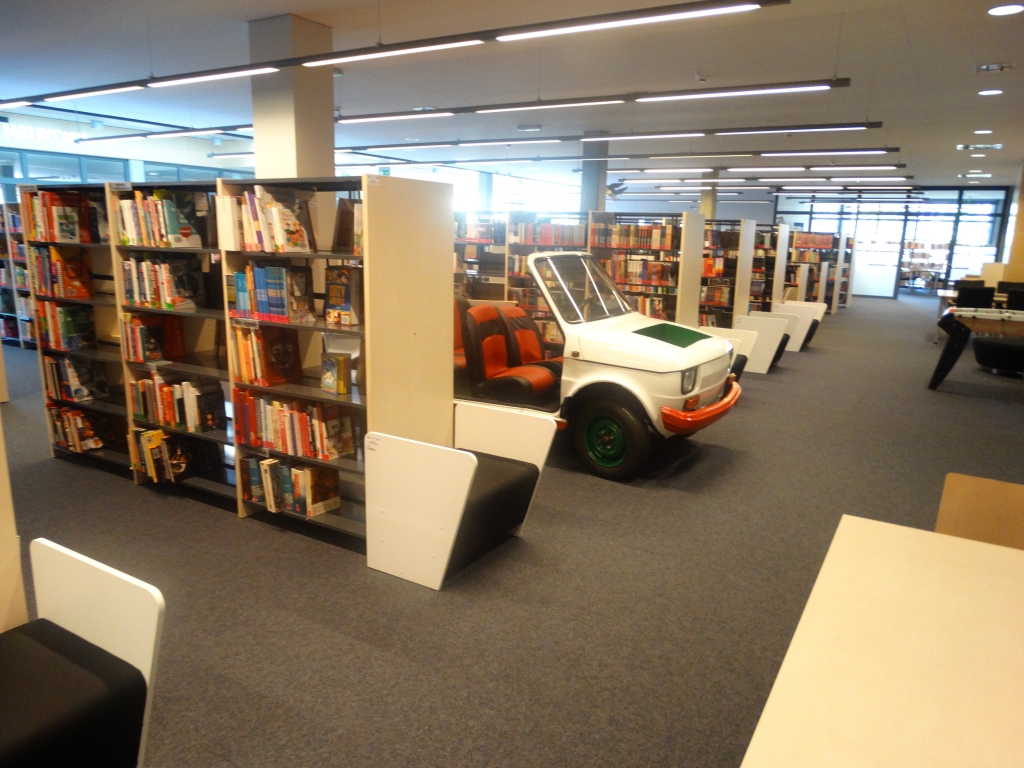 Pomieszczenie biblioteki z wieloma regałami na których są poukładane książki. W głębi samochód marki fiat, popularny "maluch" stanowiący miejsce wypoczynku i relaksu dla najmłodszych czytelników