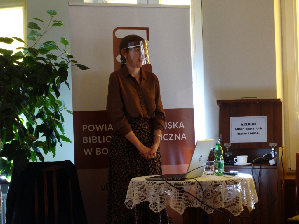 Stojąca przed małym stolikiem Dominika Drobiazg. w tle baner z logo biblioteki