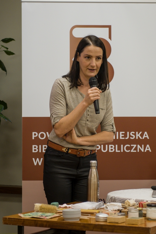 Anna Jaklewicz trzymająca w ręce mikrofon, przed nią stolik, na którym znajdują się przedmioty codziennego użytku wykonane z ekologicznych materiałów