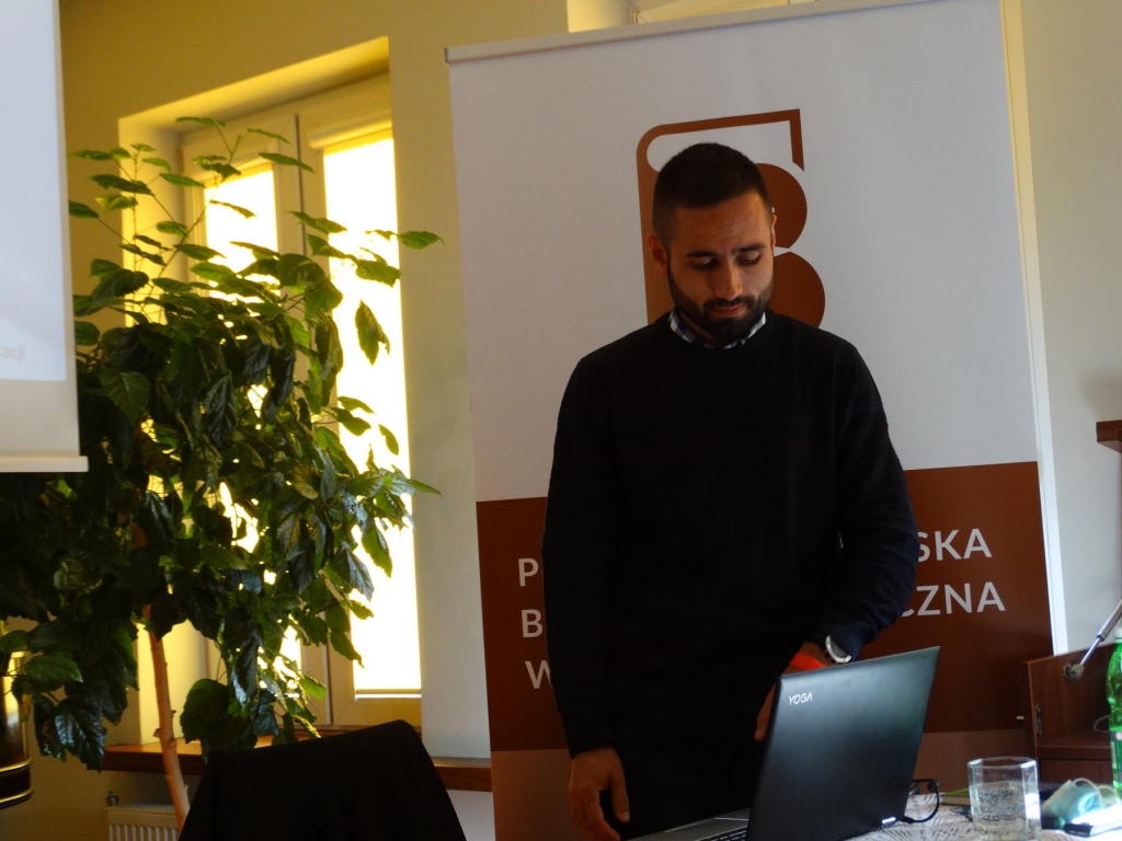 Stojący i spoglądający w ekran laptopa Michał Szmaj na tle okna i banera z logo biblioteki