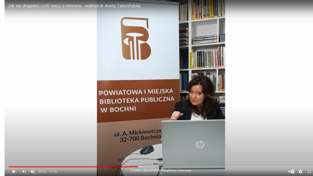 Dr hab. Aneta Załazińska spoglądająca w ekran laptopa na tle banera z logo i adresem bocheńskiej biblioteki