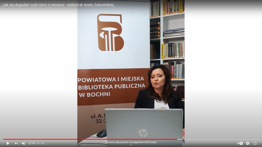 Dr hab. Aneta Załazińska spoglądająca w ekran laptopa na tle banera z logo biblioteki i regału z książkami
