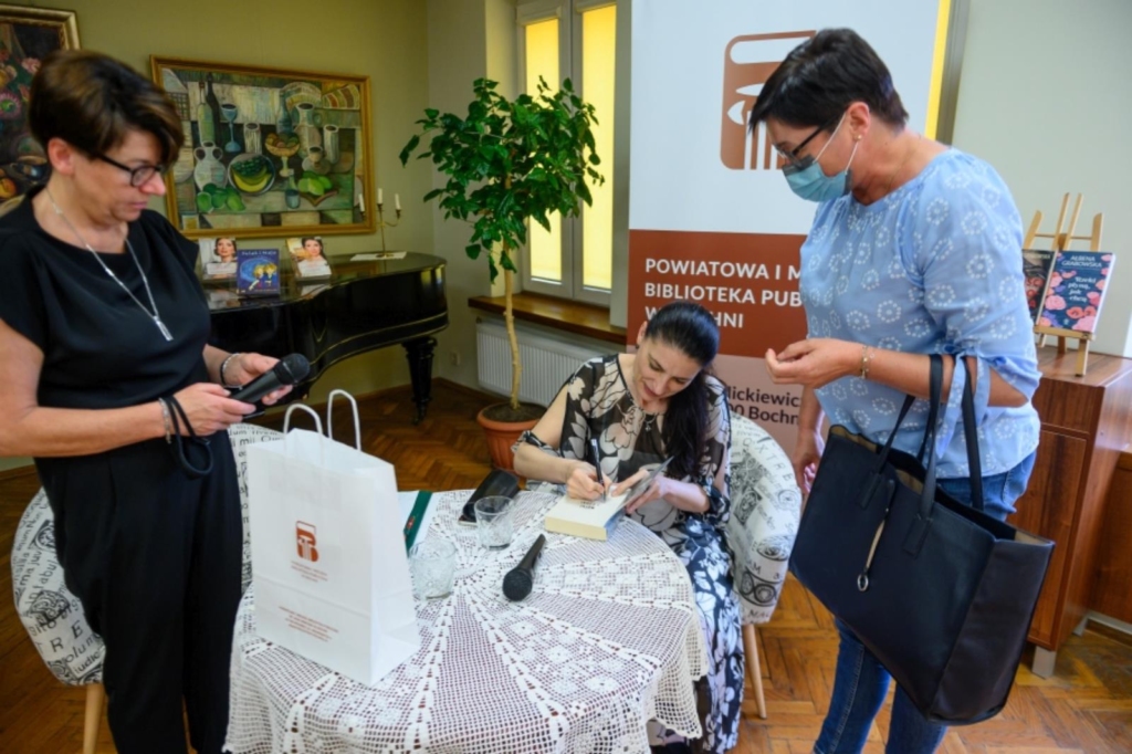 Dyrektor Biblioteki Dorota Rzepka oraz siedzaca przy stoliku Ałbena Grabowska która skjłada autograf w swojej książce, obok stoi młoda kobieta